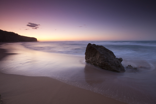 View of a large rock on the sandy shore of Twelve Apostles Marine National Park during a purple sunset with waves washing over the beach in Australia. View of a large rock on the sandy shore of Twelve Apostles Marine National Park during a purple sunset with waves washing over the beach in Australia.