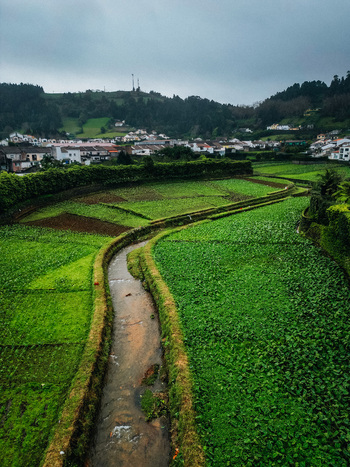 Aerial view of a meandering stream cutting through vibrant green fields towards the quaint village nestled beneath the hills Furnas Azores Portugal.