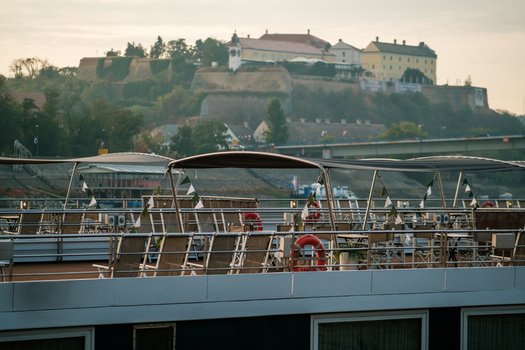 View of the deck of a river cruise ship with lounge chairs and flags with the historic Petrovaradin Fortress visible in the background in Novi Sad Vojvodina Serbia. View of the deck of a river cruise ship with lounge chairs and flags with the historic Petrovaradin Fortress visible in the background in Novi Sad Vojvodina Serbia.