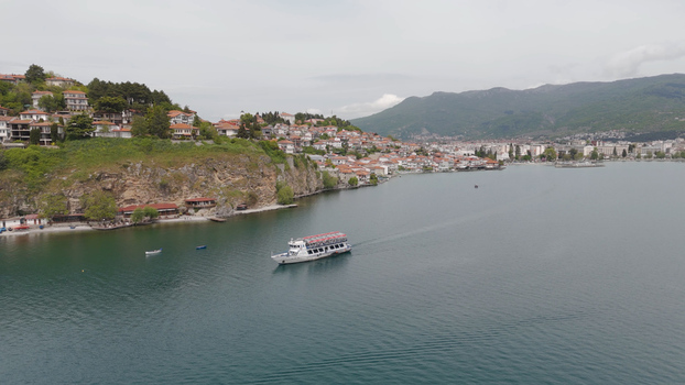 Aerial view of a passenger boat cruising on Lake Ohrid with the historic old town buildings perched on the rocky cliffside in Ohrid Municipality of Ohrid North Macedonia. Aerial view of a passenger boat cruising on Lake Ohrid with the historic old town buildings perched on the rocky cliffside in Ohrid Municipality of Ohrid North Macedonia.