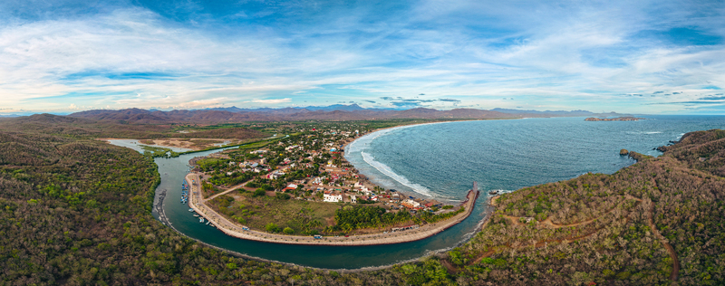 Aerial view of Playa Punta Perula showing the coastal town the curved beach the river meeting the sea and the surrounding lush landscape in Perula Jalisco Mexico. Aerial view of Playa Punta Perula showing the coastal town the curved beach the river meeting the sea and the surrounding lush landscape in Perula Jalisco Mexico.