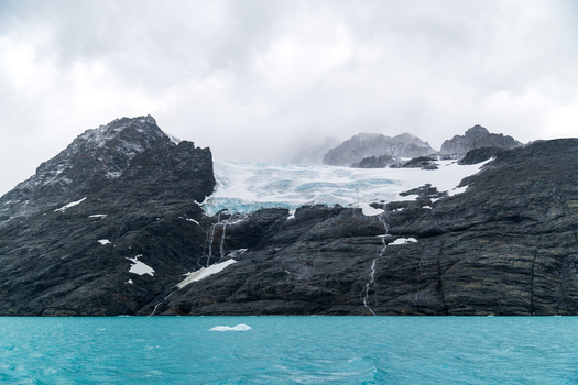 View of the glacier flowing into the turquoise waters of Drygalski Fjord surrounded by rugged dark mountain peaks under a cloudy sky in South Georgia and the South Sandwich Islands.