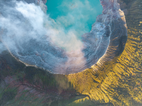 Aerial view of Ijen crater lake with vibrant turquoise water surrounded by volcanic crater rims rising steam and rugged terrain in golden hour light in Ijen East Java Indonesia.