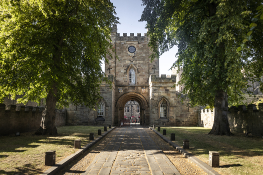 View of the historic stone gatehouse of Durham Castle with a cobblestone path leading through the archway framed by large green trees in Durham England United Kingdom. View of the historic stone gatehouse of Durham Castle with a cobblestone path leading through the archway framed by large green trees in Durham England United Kingdom.