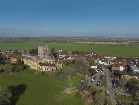 Aerial view of Tewkesbury Abbey surrounded by historic town buildings green fields and a parking area under a clear blue sky in Tewkesbury England United Kingdom.