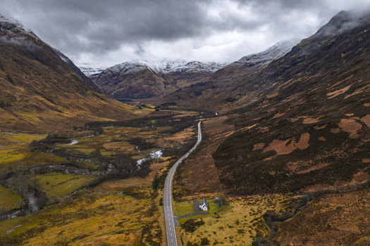 Aerial view of the winding A82 road through the dramatic Glencoe valley with snow capped mountains and a small white cottage under a moody sky Scotland United Kingdom. Aerial view of the winding A82 road through the dramatic Glencoe valley with snow capped mountains and a small white cottage under a moody sky Scotland United Kingdom.