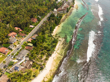 Aerial view of Thalpe Beach featuring unique rock pools turquoise ocean water lush palm trees and a coastal road lined with buildings in Talpe Southern Province Sri Lanka. Aerial view of Thalpe Beach featuring unique rock pools turquoise ocean water lush palm trees and a coastal road lined with buildings in Talpe Southern Province Sri Lanka.