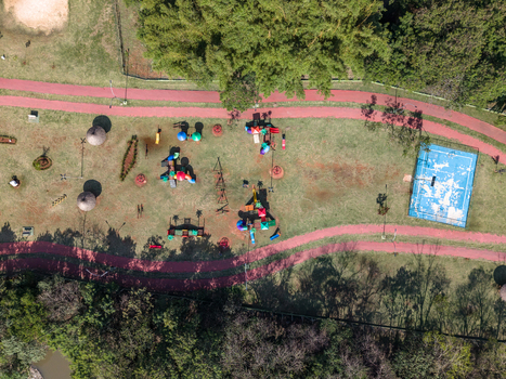Aerial view of a public park featuring a colorful playground a blue basketball court and a red running track surrounded by lush green trees in Brazil.