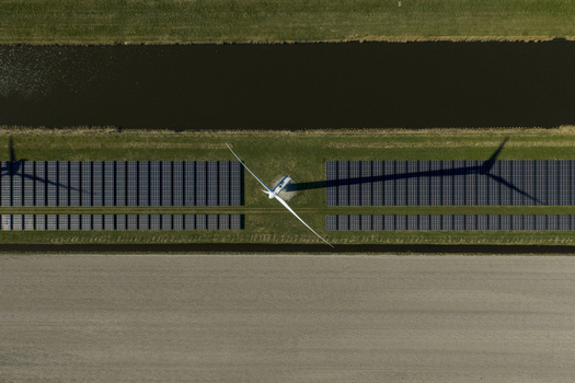 Aerial view of a wind turbine casting a long shadow over rows of solar panels next to a canal and agricultural fields in the Netherlands. Aerial view of a wind turbine casting a long shadow over rows of solar panels next to a canal and agricultural fields in the Netherlands.