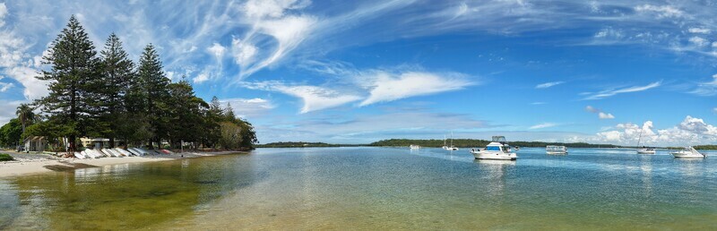 Aerial view of boats bobbing gently on the water near a sandy beach lined with lush green trees under a sky streaked with wispy clouds Noosa Heads Queensland Australia.
