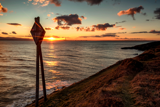 View of a wooden navigation marker on a grassy cliff edge overlooking the sea during a golden sunset in Howth Ireland. View of a wooden navigation marker on a grassy cliff edge overlooking the sea during a golden sunset in Howth Ireland.