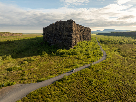 Aerial view of the massive rock formation Eyjan in the Asbyrgi canyon with a winding road and a car driving through the lush green landscape Nordurthing Iceland.