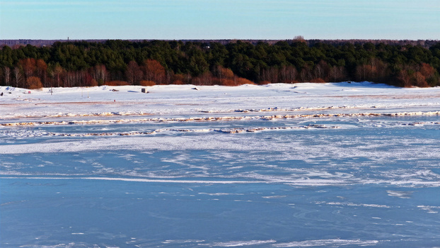 Aerial view of a frozen snow covered coastal landscape with a dense pine forest bordering the icy shoreline under a clear blue sky in Latvia. Aerial view of a frozen snow covered coastal landscape with a dense pine forest bordering the icy shoreline under a clear blue sky in Latvia.