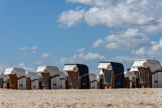 View of traditional hooded beach chairs lined up on the sandy shore of Strand Schillig under a bright blue sky with fluffy white clouds Wangerland Niedersachsen Germany. View of traditional hooded beach chairs lined up on the sandy shore of Strand Schillig under a bright blue sky with fluffy white clouds Wangerland Niedersachsen Germany.