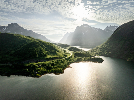 Aerial view of the Lofoten landscape featuring lush green mountains a winding road along the fjord and sunlit water under a bright summer sky in Lofoten Norway.