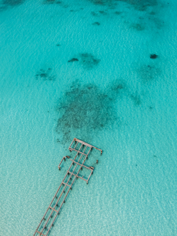 Aerial view of a weathered pier stretches into the turquoise sea revealing the sandy seabed and coral reefs beneath Vashafaru Island Maldives.