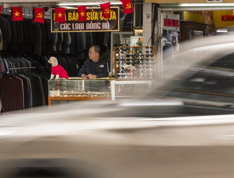 Hanoi Vietnam 11 November 2025 View of a man sitting in a watch repair shop with a small dog wearing a red shirt with a blurred vehicle passing by in the foreground.