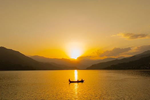 Aerial view of a small boat with people on a calm lake during golden hour surrounded by silhouetted mountains under a warm sunset sky in Nepal.