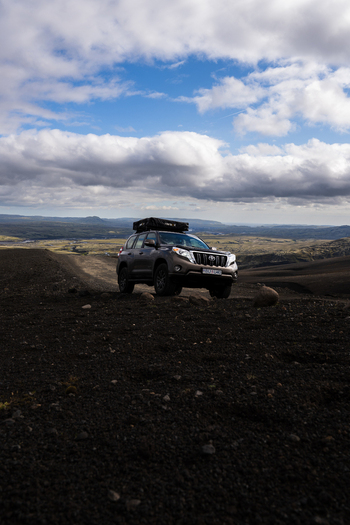 View of a dark SUV equipped for overlanding with a rooftop tent stands out against the stark black volcanic landscape under a partly cloudy sky Skaftárhreppur Iceland. View of a dark SUV equipped for overlanding with a rooftop tent stands out against the stark black volcanic landscape under a partly cloudy sky Skaftárhreppur Iceland.