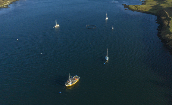Aerial view of sailboats and a yellow fishing boat anchored in the deep blue waters of Loch Dunvegan surrounded by grassy hills in Dunvegan Scotland United Kingdom. Aerial view of sailboats and a yellow fishing boat anchored in the deep blue waters of Loch Dunvegan surrounded by grassy hills in Dunvegan Scotland United Kingdom.