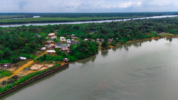 Aerial view of a small riverine village nestled along the banks of a wide river surrounded by lush tropical vegetation and palm trees in Minima Rivers Nigeria.