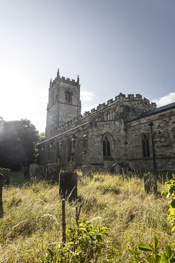 View of the historic stone church with a tall tower and crenellated walls surrounded by an old graveyard with weathered tombstones in Durham England United Kingdom. View of the historic stone church with a tall tower and crenellated walls surrounded by an old graveyard with weathered tombstones in Durham England United Kingdom.