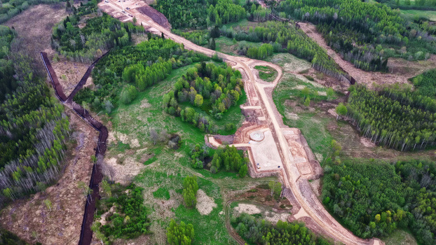 Aerial view of a dirt access road under construction winding through a lush green forest prepared for wind turbine installation in Latvia. Aerial view of a dirt access road under construction winding through a lush green forest prepared for wind turbine installation in Latvia.