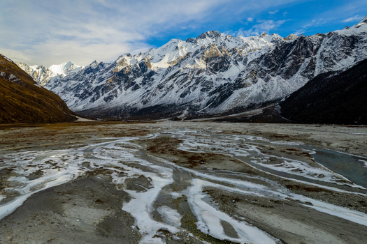 Aerial view of snow capped Himalayan peaks towering over a frozen riverbed and rugged valley floor under a clear blue sky in Nepal.