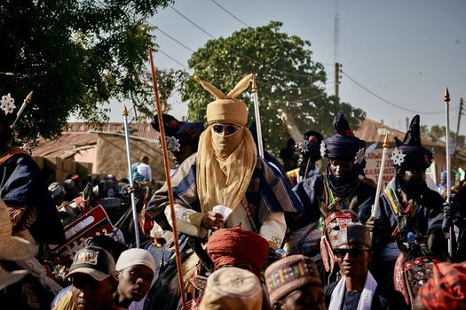 Zaria city Nigeria 21 March 2026 View of a central figure adorned in vibrant gold and blue textiles sunglasses glinting leading a procession amidst a crowd under the bright sky. Zaria city Nigeria 21 March 2026 View of a central figure adorned in vibrant gold and blue textiles sunglasses glinting leading a procession amidst a crowd under the bright sky.