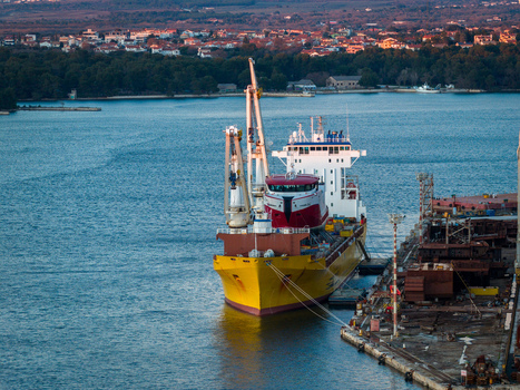 Aerial view of Uljanik Shipyard in Pula featuring a large yellow cargo ship docked at the pier with industrial cranes and the coastal city in the background Pula Croatia. Aerial view of Uljanik Shipyard in Pula featuring a large yellow cargo ship docked at the pier with industrial cranes and the coastal city in the background Pula Croatia.