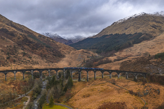Aerial view of the Glenfinnan Viaduct with a train crossing the curved stone arches surrounded by rugged hills and snow capped mountains Glenfinnan Scotland United Kingdom.