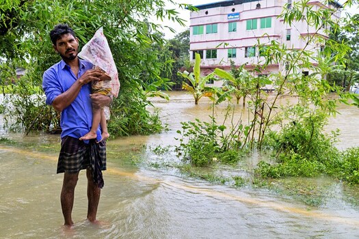 Banshkhali Upazila Chittagong Division Bangladesh 10 July 2026 View of a man carrying a child through floodwaters in front of a partially submerged building. Banshkhali Upazila Chittagong Division Bangladesh 10 July 2026 View of a man carrying a child through floodwaters in front of a partially submerged building.