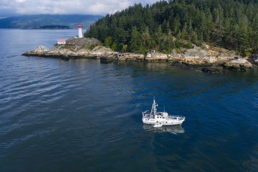 Aerial view of Point Atkinson Lighthouse situated on a rocky point surrounded by dense evergreen forest and blue waters with a fishing boat nearby in West Vancouver British Columbia Canada.