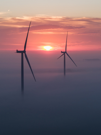 Aerial view of two wind turbines emerging from a dense layer of morning mist under a vibrant orange sunrise sky in Vrouwenpolder Zeeland Netherlands.
