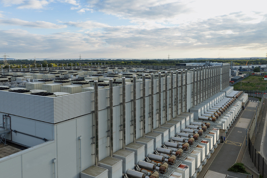 Aerial view of a large data center facility featuring extensive rooftop cooling systems industrial ventilation pipes and a perimeter fence in the United Kingdom.