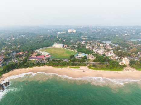 Aerial view of a sandy beach with turquoise waves a green sports stadium and lush tropical trees in Galle Southern Province Sri Lanka.