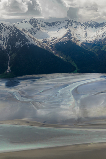 Aerial view of Alaska Wilderness featuring snow capped peaks dark evergreen forests and a vast glacial river plain with intricate silt patterns in Girdwood Alaska United States. Aerial view of Alaska Wilderness featuring snow capped peaks dark evergreen forests and a vast glacial river plain with intricate silt patterns in Girdwood Alaska United States.