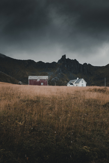 View of a red barn and a white house situated in a dry grassy field beneath dramatic dark mountains and a stormy sky in Napp Nordland Norway. View of a red barn and a white house situated in a dry grassy field beneath dramatic dark mountains and a stormy sky in Napp Nordland Norway.