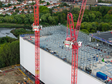 Aerial view of a data center construction site with two red tower cranes exposed steel framework and white wall panels surrounded by trees in the United Kingdom.