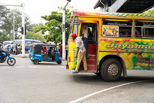 Sri Lanka 07 January 2026 View of a colorful public bus and a blue tuk tuk on a busy street with people boarding near an overpass under bright daylight.
