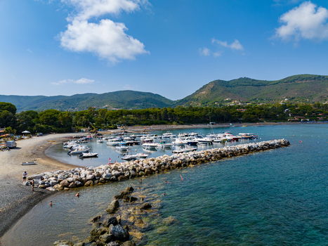 Aerial view of the marina in Lacona with numerous small boats moored behind a stone breakwater surrounded by lush green hills and a clear blue sky in Lacona Tuscany Italy.