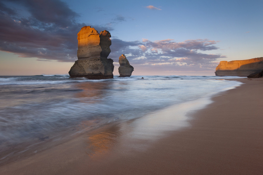 View of the Twelve Apostles limestone stacks on a sandy beach with waves coastal cliffs and golden light under a cloudy sky in Australia. View of the Twelve Apostles limestone stacks on a sandy beach with waves coastal cliffs and golden light under a cloudy sky in Australia.