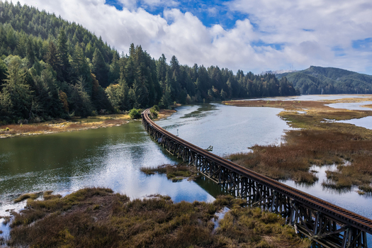 Aerial view of the Coos Bay Rail Line winding across wetlands near the Siuslaw River in Florence Oregon with calm water marshland and evergreen forest.