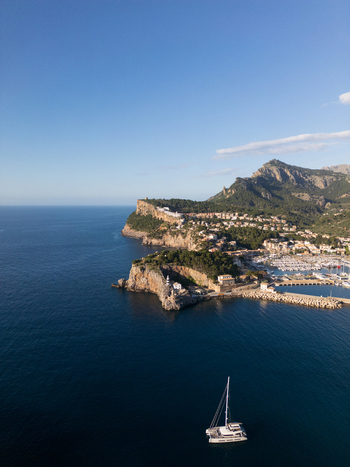 Aerial view of the harbor with a lighthouse on the rocky cliffs and a catamaran sailing on the blue Mediterranean sea in Port de Soller Balearic Islands Spain.