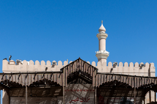 View of the minaret of a mosque rising above a traditional structure with a wooden roof against a clear blue sky in Muscat Muscat Governorate Oman. View of the minaret of a mosque rising above a traditional structure with a wooden roof against a clear blue sky in Muscat Muscat Governorate Oman.