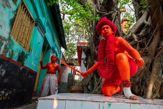 Munshiganj Bangladesh 13 April 2026 View of performers with orange body paint and traditional costumes during a local festival in front of a turquoise building and ancient tree.