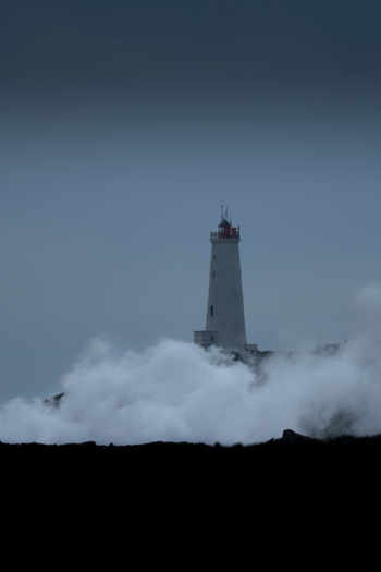 View of Reykjanes Lighthouse rising above the crashing sea spray and dark volcanic rocks under a moody overcast sky in Keflavik Reykjanesbaer Iceland.