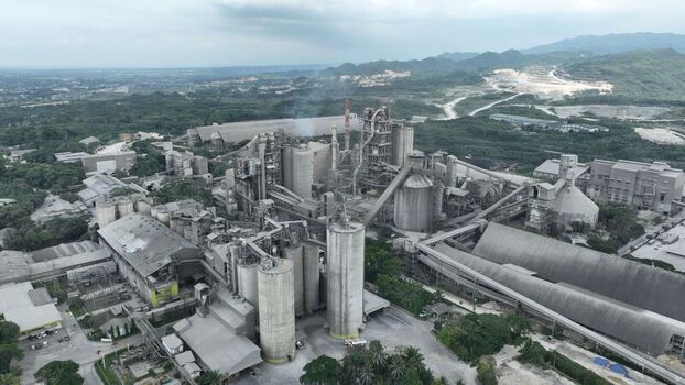 Aerial view of a large industrial cement factory with tall silos processing structures and conveyor belts surrounded by green hills in Klapanunggal West Java Indonesia. Aerial view of a large industrial cement factory with tall silos processing structures and conveyor belts surrounded by green hills in Klapanunggal West Java Indonesia.
