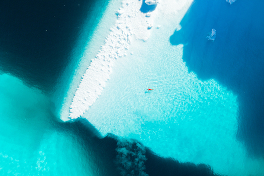Aerial view of a person swimming in clear turquoise water next to a large white iceberg in Greenland. Aerial view of a person swimming in clear turquoise water next to a large white iceberg in Greenland.
