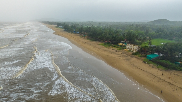 Aerial view of Gokarna beach with waves crashing on the sandy shore lush palm trees and coastal buildings under a cloudy sky in Gokarna Karnataka India.